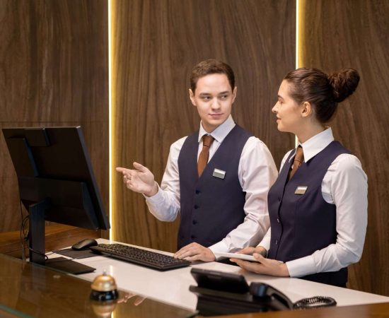 one-two-young-hotel-receptionists-standing-by-counter-looking-colleague-with-touchpad-pointing-computer-screen (1) (1) (1)